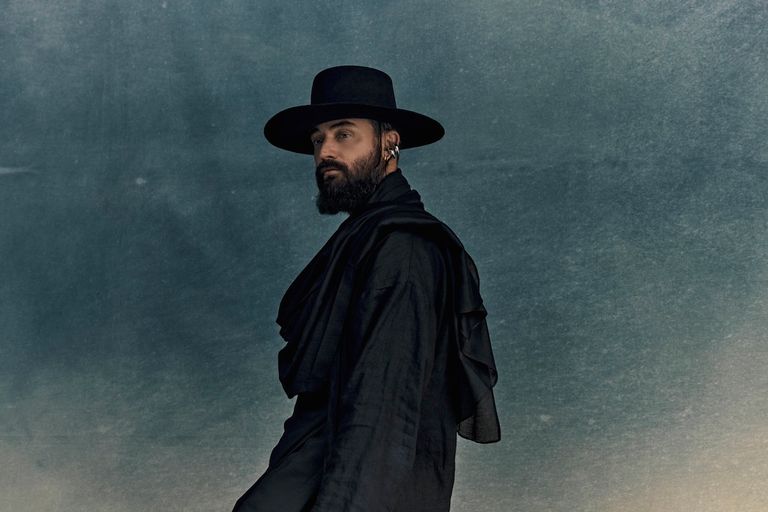 Musician RY X posing in profile against a textured blue backdrop, wearing a wide-brimmed black hat, dark draped clothing, and a full beard.